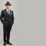 Dapper 1930s gentleman wearing tailored charcoal suit with natural shoulders, crisp white spread-collar shirt, silk tie, fedora hat, and polished oxford shoes standing confidently in classic menswear pose