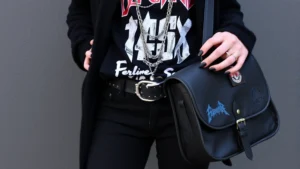 Person wearing black skinny jeans, oversized band graphic t-shirt, open black cardigan, studded belt, multiple layered silver chains, black nail polish, holding vintage messenger bag with band patches
