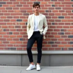 Young man wearing oversized cream blazer with tapered black trousers and white sneakers, standing confidently against urban brick wall, natural daylight, modern minimalist aesthetic