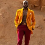 Man wearing mustard-yellow velvet blazer with wide lapels, burgundy bell-bottom trousers, and gold chain necklace, standing confidently in natural lighting with earth-tone background