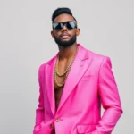 Man wearing oversized neon pink blazer with geometric patterns, bold sunglasses, and chunky gold chain necklace, confident pose against neutral backdrop, professional studio lighting