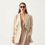 Professional fashion model wearing oversized neutral blazer, minimalist slip dress, and tiny rectangular sunglasses, standing against clean white backdrop, soft natural lighting, high-fashion editorial style