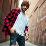 Male model wearing oversized flannel shirt in red and black plaid, distressed dark jeans, white vintage band t-shirt layered underneath, white canvas sneakers, brown knit beanie, standing in urban alleyway with brick wall background, natural overcast daylight, candid relaxed pose