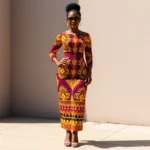 Woman confidently wearing vibrant Ankara dress with bold geometric patterns, standing in natural sunlight, professional styling with structured accessories and neutral footwear, sophisticated posture