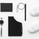 Minimalist fashion flat lay featuring black and white clothing pieces, silver watch, and clean white sneakers arranged on light background with architectural precision and negative space