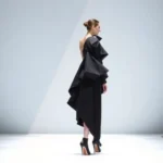 Asymmetrical black avant-garde garment with exaggerated shoulder structure, worn on minimalist white runway, dramatic side-lighting creating sharp shadows, model in profile showcasing geometric silhouette