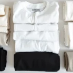 Flat lay of minimalist wardrobe basics: neutral folded clothing, quality fabrics, simple white and black pieces arranged neatly