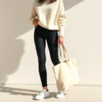 Woman wearing cream oversized sweater, fitted dark jeans, white leather sneakers, holding canvas tote bag, standing in natural sunlight, minimalist aesthetic, relaxed confident posture