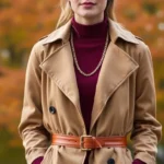 Woman wearing rich burgundy cashmere sweater layered under camel trench coat, styled with cognac leather belt and gold chain necklace, standing against autumn foliage backdrop