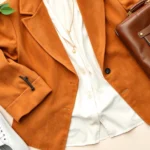 Styled flat lay featuring a 1970s suede blazer draped over contemporary white linen, paired with modern minimalist gold jewelry, sleek white sneakers, and a structured contemporary leather bag on light neutral background