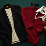 Overhead flat lay of dark academia wardrobe essentials: cream cable-knit sweater, black blazer, burgundy cardigan, white collared shirt, arranged artfully on deep forest green background with leather satchel and vintage eyeglasses