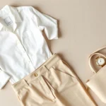 Overhead flat lay of minimalist fashion essentials: white button-up, tailored beige trousers, simple gold watch, and cream leather handbag arranged on neutral background with soft natural lighting