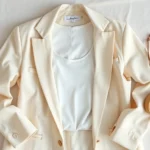 Overhead flat lay of minimalist clothing items arranged aesthetically: cream linen blazer, white tank top, natural fabric textures, subtle color palette, professional styling, bright natural lighting, high-quality fashion photography