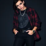 Person wearing black skinny jeans, layered band t-shirt under oversized plaid button-up, silver chain accessories, dramatic dark eye makeup with black eyeliner wings, side-swept black hair with colored streaks, standing confidently against moody backdrop