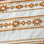 Close-up of intricate hand-woven geometric patterns on white cotton fabric, showing detailed cross-stitch embroidery with traditional Ethiopian design motifs in earth tones, natural lighting highlighting textile texture