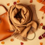 Overhead flat lay of fall fashion accessories: cognac leather structured handbag, oversized camel wool scarf, gold layered chain necklaces, burgundy leather statement belt with gold buckle, arranged on warm neutral background with autumn leaves scattered artfully, professional product photography style, natural daylight
