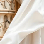 Close-up of flowing white linen chiton fabric with careful pleats and folds, draped elegantly on a classical marble column, showing the texture and weave of ancient Greek textile construction, soft natural lighting