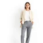 A minimalist woman wearing a perfectly coordinated neutral outfit consisting of cream linen blazer, white t-shirt, and tailored gray trousers, standing against a clean white backdrop, embodying capsule wardrobe elegance and intentional style