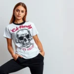 Young woman wearing vibrant Ed Hardy graphic tee with skull motif and rhinestone details, styled with black tailored trousers and minimalist white sneakers, confident pose against neutral background, natural lighting, contemporary fashion photography, modern interpretation of early 2000s luxury aesthetic