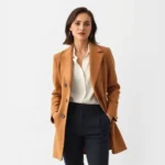 Elegant woman wearing tailored camel coat, white silk blouse, and dark trousers, standing against minimalist white background, exuding sophisticated confidence and timeless style