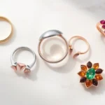 Flat lay photography of five diverse fashion rings in different styles including minimalist gold band, chunky silver signet ring, stacked rose gold rings, colorful gemstone cocktail ring, and nature-inspired botanical design ring arranged on white marble surface with soft natural lighting