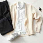 Minimalist fashion flat lay featuring neutral-toned basics: white linen shirt, cream cardigan, black tailored trousers, white leather sneakers, and small accessories arranged artfully on light marble surface with natural window light