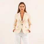 Woman wearing tailored cream blazer and fitted white trousers, standing confidently against neutral backdrop, professional polished appearance, natural lighting, fashion editorial style