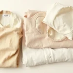 Minimalist fashion flat lay featuring neutral linen and cotton garments, cream and taupe folded clothing, natural textures, warm studio lighting, no text or logos visible, photorealistic styling