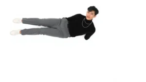 Asian male model wearing perfectly fitted black turtleneck tucked into high-waisted gray tailored trousers, cropped to ankle length, paired with white minimalist leather sneakers and delicate silver chain necklace, standing against neutral white background in confident pose