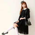 A young woman wearing a Classic Lolita coordinate with a black JSK dress, white knee-high socks, black Mary Jane shoes, an ornate headbow, and a structured lace bag, posing elegantly in soft natural lighting with a neutral background.