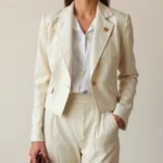 Woman wearing a high-waisted cream linen blazer over a white silk button-down shirt tucked into vintage-inspired tailored trousers, styled with a delicate gold brooch and holding a structured leather handbag, photographed against a soft neutral background