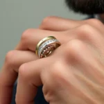 Close-up of three stacked men's fashion rings in different metals—gold, silver, and rose gold—on a man's hand, showing intricate band details and textures against neutral background