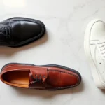 Professional overhead flat lay of three premium men's dress shoes in black oxford, brown loafer, and white leather sneaker, arranged on marble surface with soft natural lighting