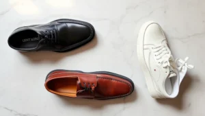 Professional overhead flat lay of three premium men's dress shoes in black oxford, brown loafer, and white leather sneaker, arranged on marble surface with soft natural lighting
