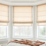 Luxurious roman shades in soft neutral tones draped elegantly on large windows, showing sophisticated fabric texture and precise pleating, natural daylight filtering through, modern minimalist bedroom setting