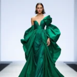 Fashion model wearing emerald green sculptural gown with dramatic draped sleeves and oversized silhouette on minimalist runway, professional lighting, haute couture aesthetic, confident pose