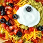 Close-up overhead shot of a colorful traditional 7-layer dip in a clear glass baking dish showing distinct layers of guacamole, sour cream, salsa, cheese, and black olives with tortilla chips arranged around the edges, warm natural lighting, vibrant colors