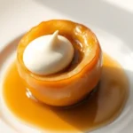 Golden-brown baked apple with caramelized filling, garnished with a single vanilla cream quenelle, served on a minimalist white ceramic plate with soft natural lighting highlighting the glossy caramel surface and deep apple tones