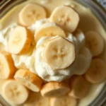 Overhead shot of vintage banana pudding in glass dish showing distinct layers of creamy custard, vanilla wafers, and fresh banana slices, soft natural lighting, classic tableware