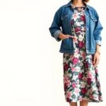 Woman wearing 1950s floral print dress styled with modern white sneakers, oversized denim jacket, and minimalist gold jewelry, standing in natural light against neutral background, fashion-forward vintage styling