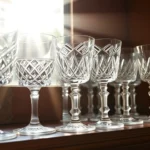 Elegant vintage cut crystal glasses arranged on antique wooden shelf, sunlight creating prismatic reflections, various geometric patterns visible, soft warm lighting, close-up detail shot