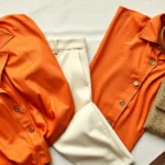 Flat lay composition of 1970s vintage clothing pieces including a burnt orange silk blouse, cream wool trousers, and a brown leather belt arranged artfully on a neutral background, natural lighting emphasizing fabric texture and quality craftsmanship