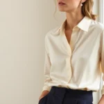 Sophisticated woman wearing classic cream silk blouse tucked into tailored navy wool trousers, standing by a window with natural light, embodying understated elegance and quiet luxury aesthetic, pearl studs visible, neutral background
