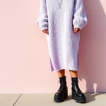 A person wearing a soft lavender oversized sweater paired with black platform boots and silver chain accessories, standing against a pale pink wall in soft natural lighting, showcasing pastel goth contrast styling