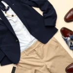 Flat lay of preppy wardrobe essentials: navy blazer, white oxford shirt, tailored khaki trousers, pearl necklace, leather loafers, and silk scarf arranged on cream background. Clean, minimalist composition showcasing classic pieces