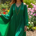 Woman wearing emerald green flowing maxi dress with gold layered necklaces, standing in spring garden with blooming flowers, natural sunlight, confident pose, minimalist styling