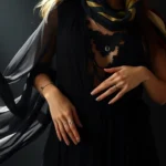 Woman wearing flowing black chiffon maxi dress with layered lace overlays, stacked silver rings, delicate chain necklaces, and a draped silk scarf in jewel tones, standing with ethereal grace in dramatic lighting