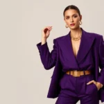 Woman wearing oversized structured blazer with pronounced shoulders in jewel-tone purple, paired with high-waisted tailored trousers, metallic gold belt, and chunky gold jewelry, confident pose against neutral background, professional studio lighting, fashion editorial style