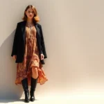 Elegant woman wearing layered vintage 1970s bohemian dress with modern minimalist black boots and structured contemporary blazer, standing in natural sunlight near neutral backdrop, showcasing era-mixing style