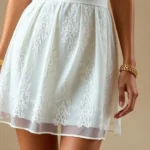 Close-up of elegant white mini dress with delicate lace detailing, styled with gold jewelry and nude heels, photographed on model in studio lighting with neutral background